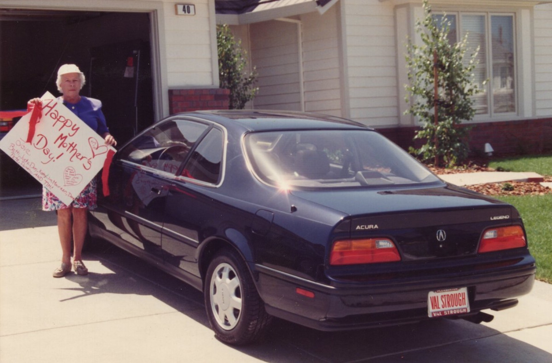 An elderly woman standing outside a house, holding a colorful sign that says 'Happy Mother's Day!' next to a dark blue Acura Legend car.