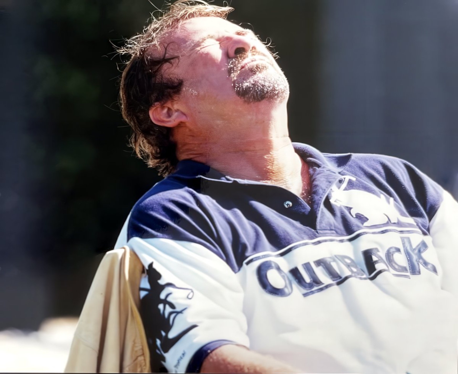 A man with medium-length hair and a beard is sitting outdoors, tilting his head back with eyes closed, enjoying the sunlight. He is wearing a blue and white polo shirt with the word 'Outback' prominently displayed.