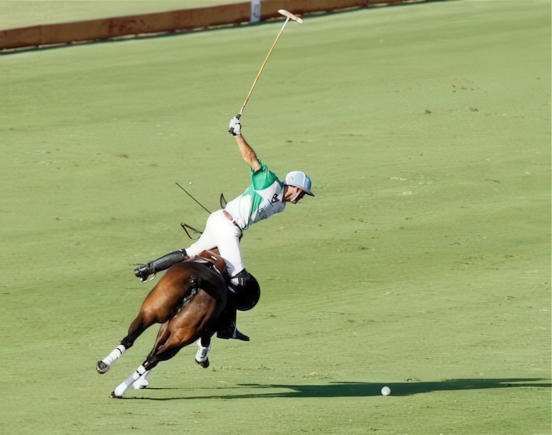 A polo player in mid-action, leaning forward on a galloping horse, swinging a mallet to strike a ball on the grass field.