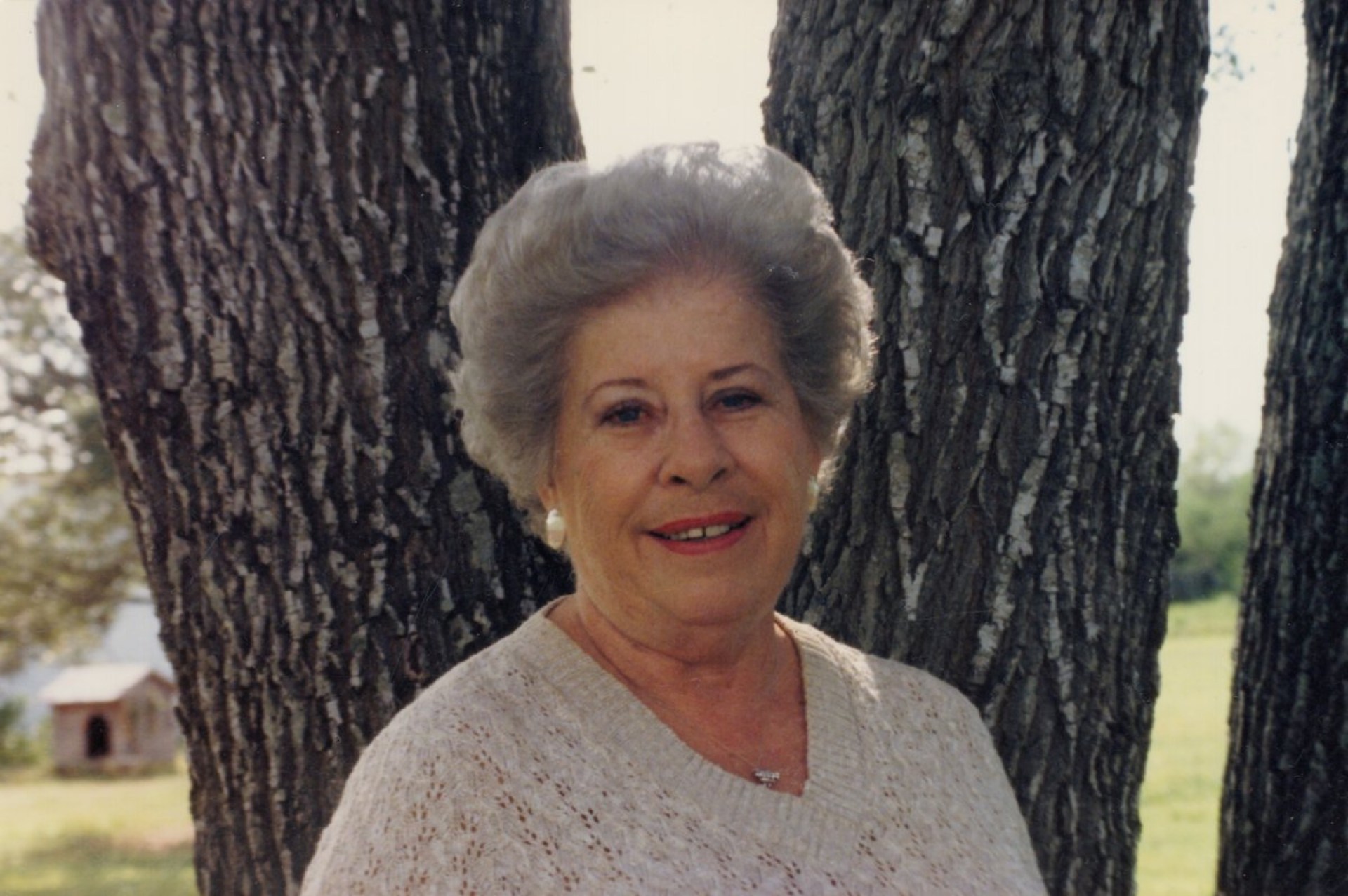 A smiling elderly woman with short gray hair wearing a light sweater, standing outdoors in front of trees.