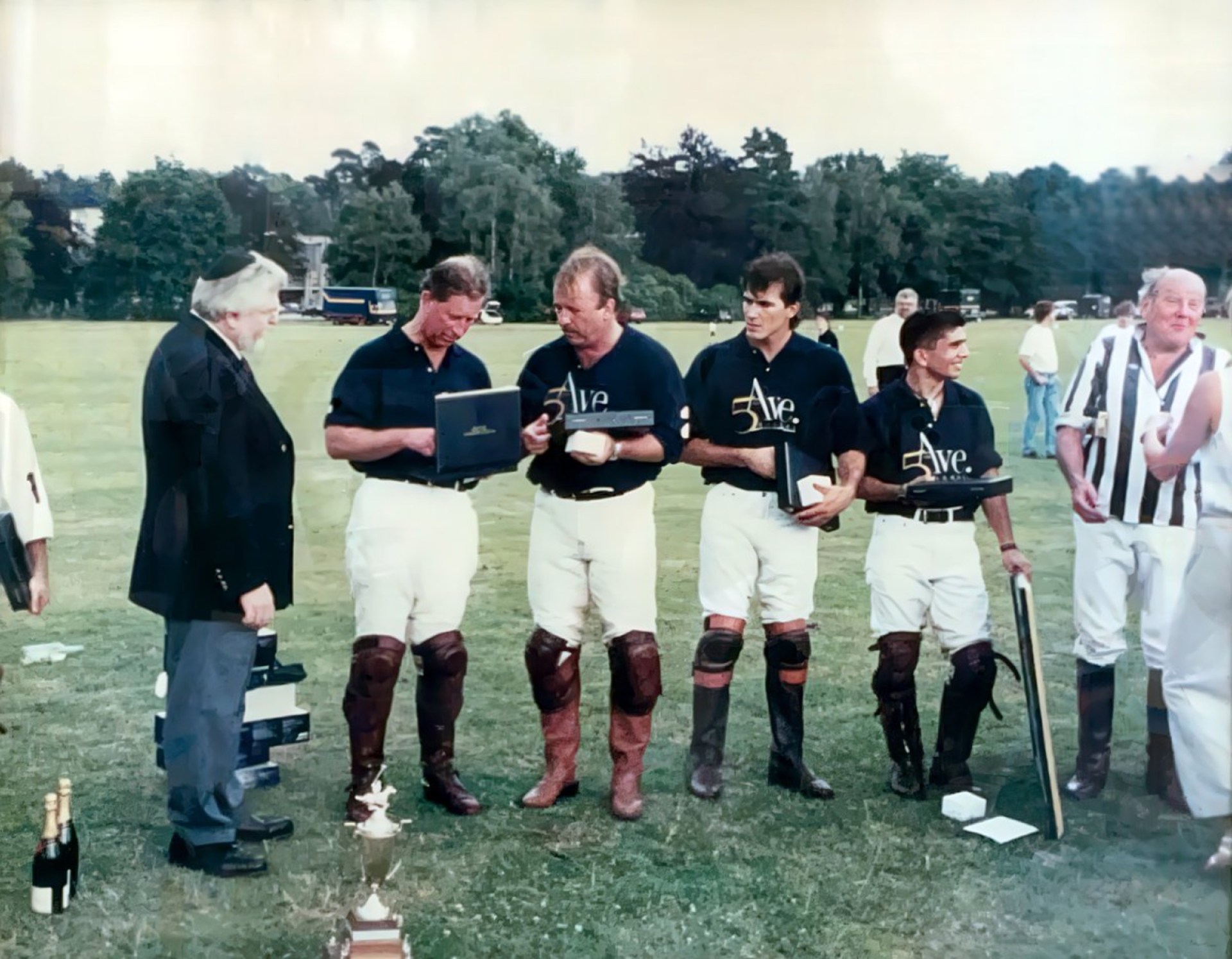 A group of five men, dressed in polo attire, stand on a grassy field with a trophy and champagne in front of them. One man is holding a plaque, while they appear to be engaged in conversation. A man in a black suit stands nearby observing, and another man in striped attire is in the background.