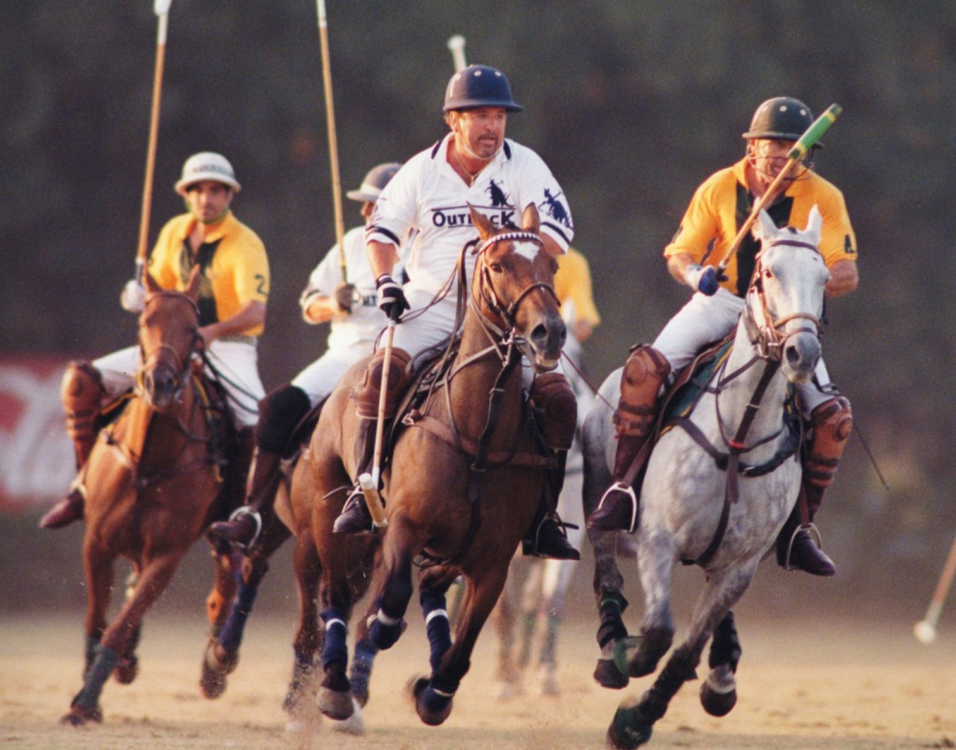 A dynamic scene of a polo match featuring players on horseback. Two riders are prominently competing, with one in a white uniform and the other in an orange uniform. The background shows additional players and horses in motion.