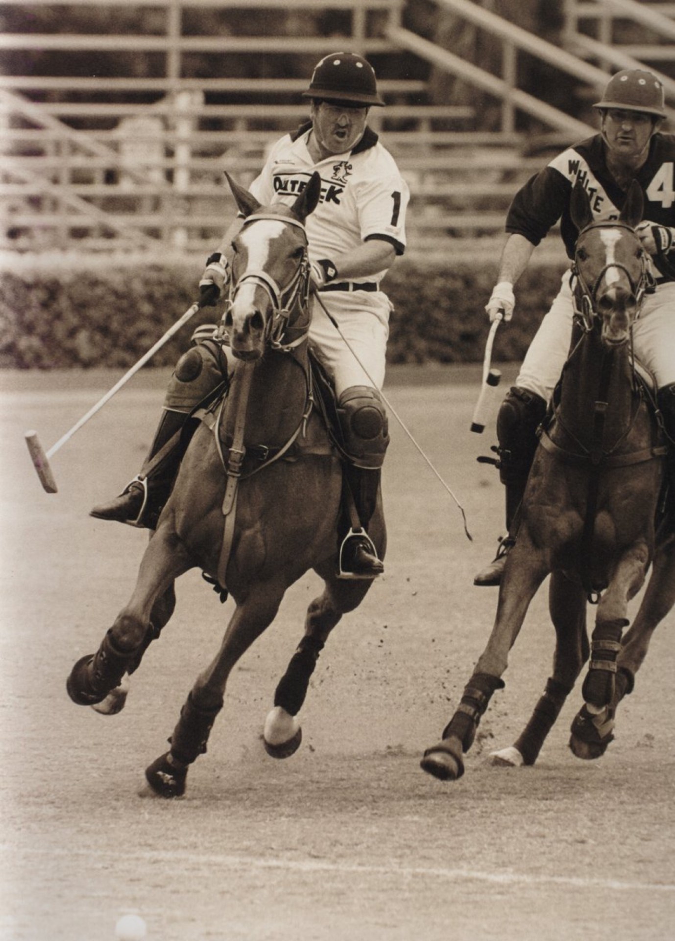 Action shot of two polo players riding horses, one wearing a white jersey and the other wearing a dark jersey, both in motion during a match.