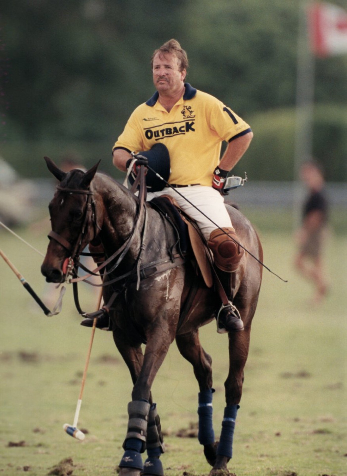 A polo player wearing a yellow Outback shirt rides a horse on a grassy field, holding a polo mallet, with players in the background.
