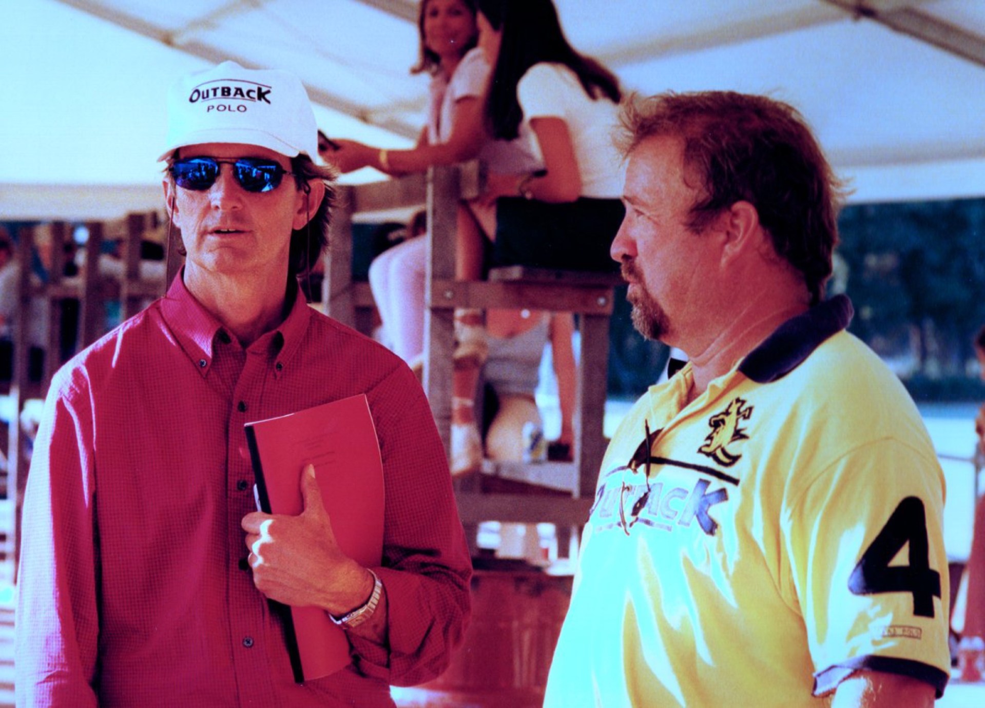 Two men conversing under a tent; one wearing a white cap and sunglasses, holding a red folder, and the other in a yellow polo shirt.